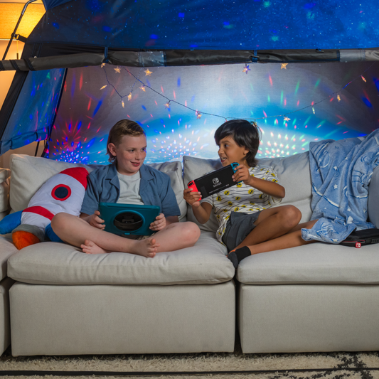 Two boys playing with handheld consoles on the living room couch with a MyFort tent set up.